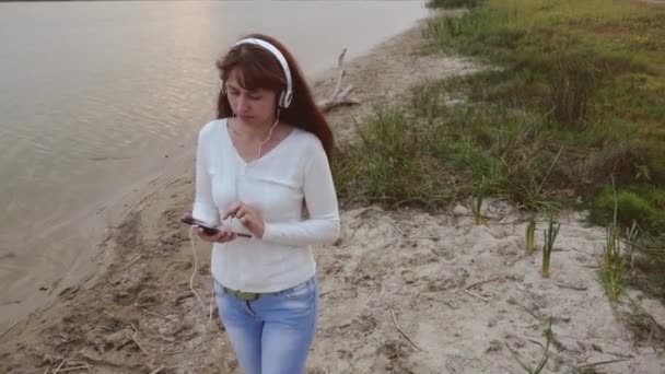 Chica con auriculares caminando sobre arena junto al agua y escuchando música en la tableta. chica camina en la playa y cheques de correo en la tableta en línea . — Vídeos de Stock