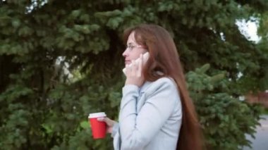 neşeli iş kadını ışık takım elbiseli bardaklarda işe gider, kız parkta kahve içer ve Smartphone'da konuşuyor