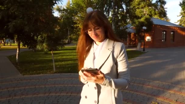 Bella donna d'affari con gli occhiali a piedi lungo il vicolo nel parco con tablet in mano, ragazza in un abito da lavoro leggero va al lavoro . — Video Stock