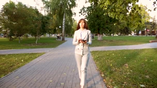 Hermosa mujer de negocios camina por el callejón en el parque con la tableta en sus manos, chica en un traje de negocios ligero va a trabajar . — Vídeos de Stock