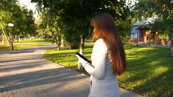 Flicka med surfplatta i ljus kostym går till jobbet. vacker affärskvinna promenader längs gränd i park med tablett i händerna. — Stockvideo