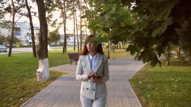 光ビジネス スーツでタブレットを持つ少女は、仕事に行きます。彼女の手のタブレットで公園の路地沿い歩く美しい女性実業家. — ストック動画