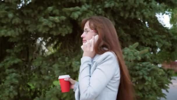 Femme d'affaires gaie dans des lunettes en costume léger va au travail, fille boit du café dans le parc et parle sur un smartphone — Video