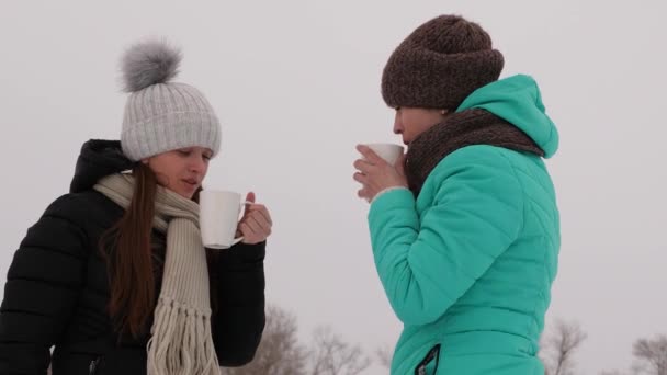 Мама и взрослая дочь смеются и улыбаются в морозный день. Красивые девушки пьют кофе из белого стекла в зимнем парке . — стоковое видео