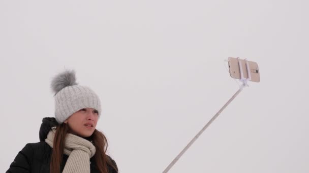 Молода дівчина фотографується на смартфоні і самосвітанку, в зимовому парку - в різдвяний ранок. Щаслива дівчина гуляє в сніжному лісі, падаючи білий сніг. крупним планом — стокове відео
