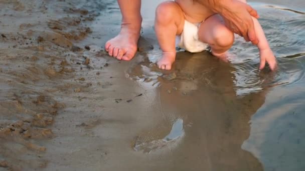 Petit bébé en couches joue sur la plage par la rivière. Le gamin se met les mains dans l'eau. Gros plan. Mouvement lent . — Video