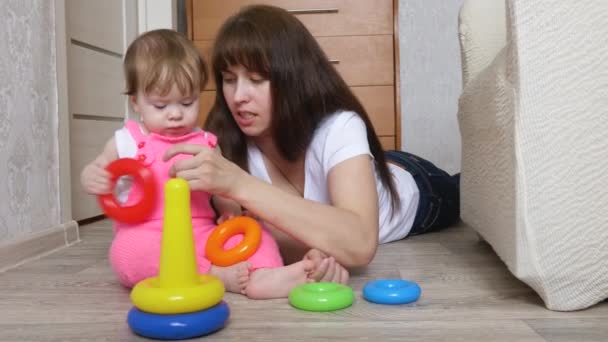 Mãe brinca com a filha pequena em anéis coloridos eles coletam pirâmide de brinquedos na sala das crianças no chão — Vídeo de Stock