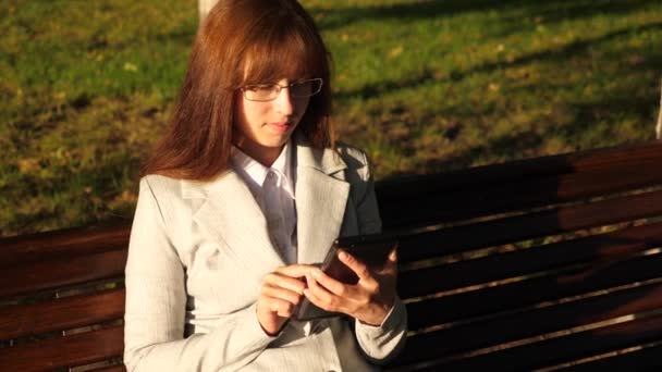 Hermosa mujer de negocios con gafas comprueba el correo electrónico en la tableta en el parque en un banco — Vídeo de stock