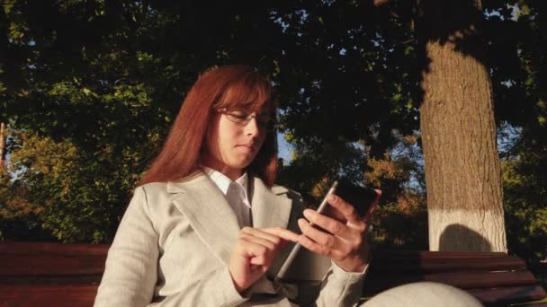 Schöne Geschäftsfrau mit Brille, die eine E-Mail auf dem Tablet im Park auf einer Bank tippt, die von der hellen Sonne beleuchtet wird — Stockvideo