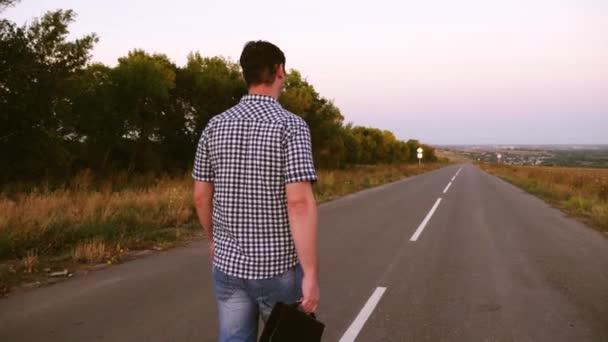 Man loopt langs een asfaltweg met een zwarte koffer in zijn hand — Stockvideo