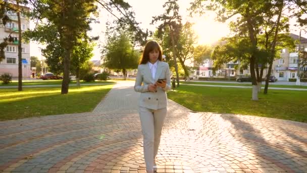 Menina em terno leve vai trabalhar com tablet na mão. Mulher bonita empresária caminha ao longo beco no parque e verifica e-mail no tablet . — Vídeo de Stock