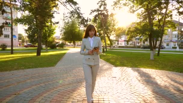 Chica en traje de luz va a trabajar con la tableta en la mano. Hermosa mujer de negocios camina por el callejón en el parque y comprueba el correo electrónico en la tableta . — Vídeo de stock