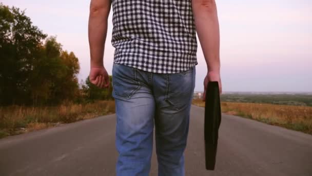 Бизнесмен в клетчатой рубашке идет по асфальтовой дороге с черным портфелем в руке. макро — стоковое видео