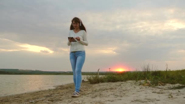 Flicka i strålar av sunset promenader på stranden och kontrollera mail på en surfplatta online. flicka med hörlurar promenader längs stranden med surfplattan och lyssna på musik. — Stockvideo