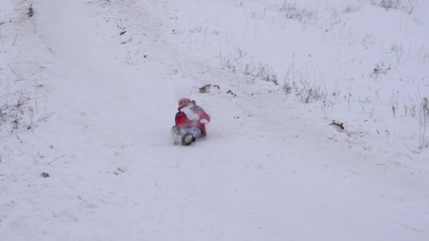 Niño feliz se desliza en el deslizamiento de nieve en trineo. La niña está jugando en invierno en el parque. Movimiento lento — Vídeos de Stock