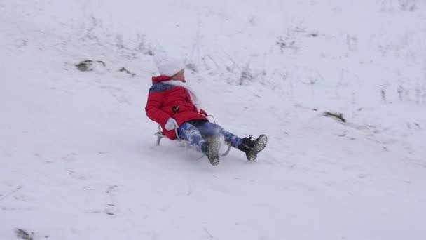 Szczęśliwe dziecko w czerwonej kurtce slajdów poprzez śnieg w sanie i śmieje się. Wesoła dziewczyna gra w święta Bożego Narodzenia w winter park. Zwolnionym tempie — Wideo stockowe