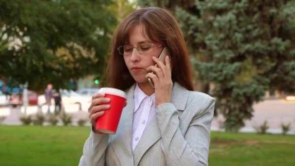 Γυναίκα των επιχειρήσεων με τα γυαλιά στο φως κοστούμι μιλώντας στο smartphone και να πίνει καφέ στο πάρκο — Αρχείο Βίντεο
