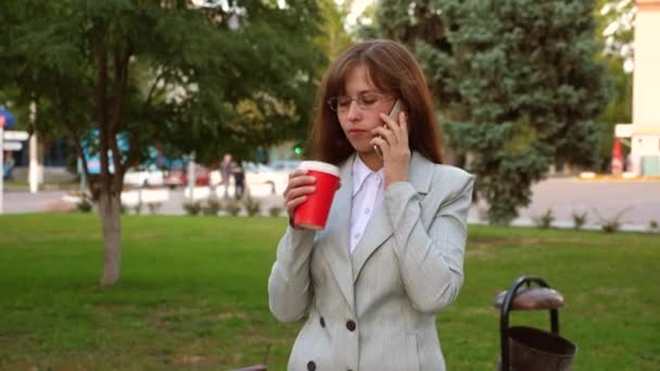 Hermosa mujer de negocios con gafas en traje ligero beber café en el parque y hablar en un teléfono inteligente — Vídeos de Stock