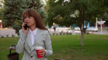 kahve içme ve Smartphone'da konuşurken Park işkadını ışık takım elbiseli gözlüklü yürür