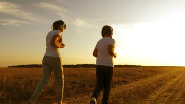 Chicas adolescentes en auriculares haciendo deportes trotando en el campo. niñas están entrenando al atardecer y escuchando música — Vídeos de Stock