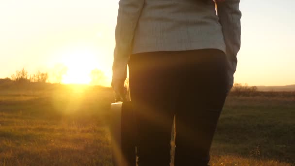 Mujer de negocios con maletín negro en la mano camina por el campo iluminado por hermosos rayos del sol. Primer plano. Movimiento lento . — Vídeos de Stock