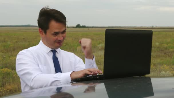 Happy businessman in blue and white shirt works in field on computer on the roof of car. — Stock Video