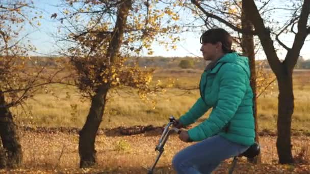 Jeune fille en veste verte fait du vélo dans le parc d'automne sur un fond d'arbres jaunes. Mouvement lent — Video
