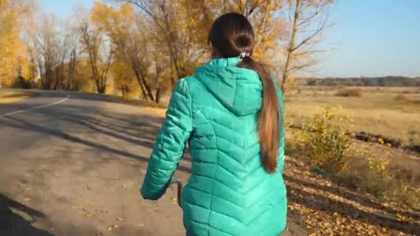 Молода дівчина в зеленій куртці їде на велосипеді по дорозі в осінньому парку, на тлі жовтих дерев . — стокове відео