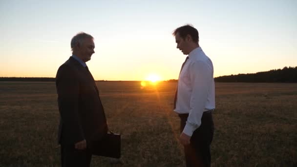 Zwei Geschäftsleute reichen einander schwarze Aktentasche und schütteln sich bei Sonnenuntergang auf dem Feld die Hand — Stockvideo