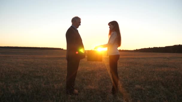 Un anciano hombre de negocios y una joven mujer de negocios hicieron un trato, se pasaron el maletín negro y se dieron la mano al atardecer en el campo. . — Vídeos de Stock