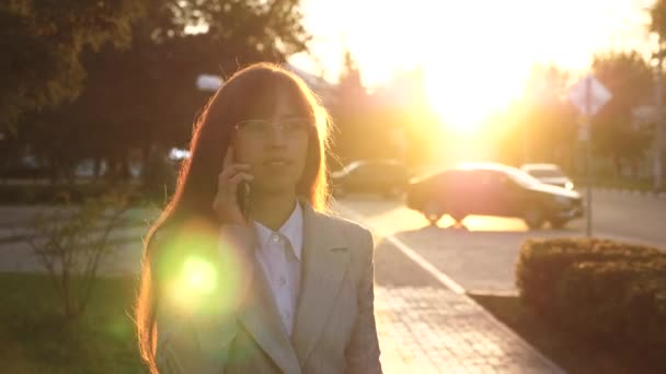 Donna d'affari con occhiali e tuta leggera che cammina per strada e parla su smartphone. bella donna a piedi lungo la strada della città nei raggi gialli del tramonto — Video Stock