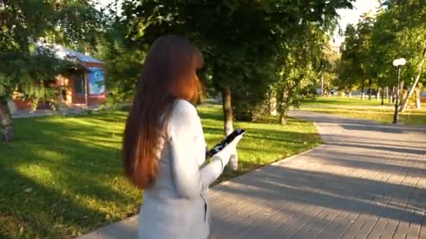 Bela mulher de negócios caminha ao longo beco no parque com tablet em suas mãos. menina com tablet em terno de negócios leve vai trabalhar . — Vídeo de Stock