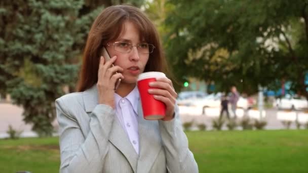 Mulher de negócios com óculos bebendo café quente no parque e falando no smartphone — Vídeo de Stock