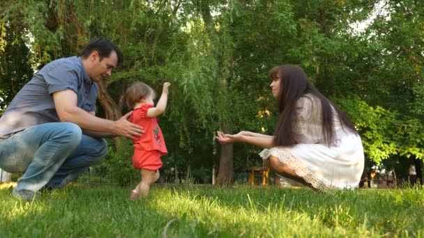 Dad and Mom learn to walk little daughter in park on green grass. Mom kisses daughter on the cheek and laughs with dad. — Stock Video