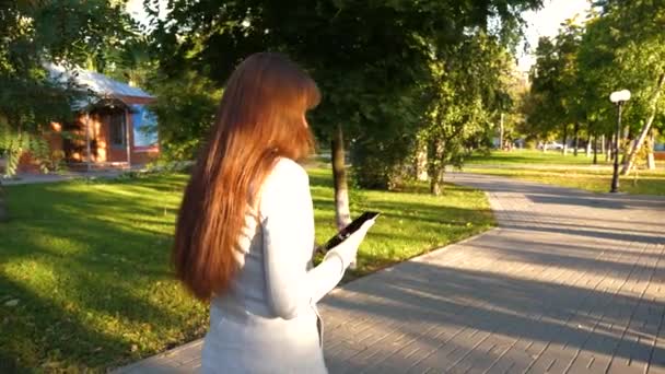 Girl with tablet in a light business suit goes to work. beautiful businesswoman walks along alley in park with tablet in her hands. — Stock Video