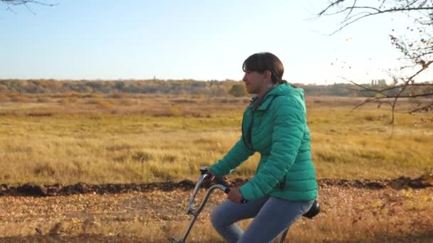 Niña en chaqueta verde paseos en bicicleta en el parque de otoño sobre un fondo de árboles amarillos. Movimiento lento — Vídeo de stock