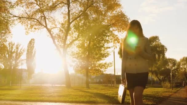 Mujer con maletín y teléfono camina por el callejón de otoño en rayos de hermoso atardecer . — Vídeos de Stock