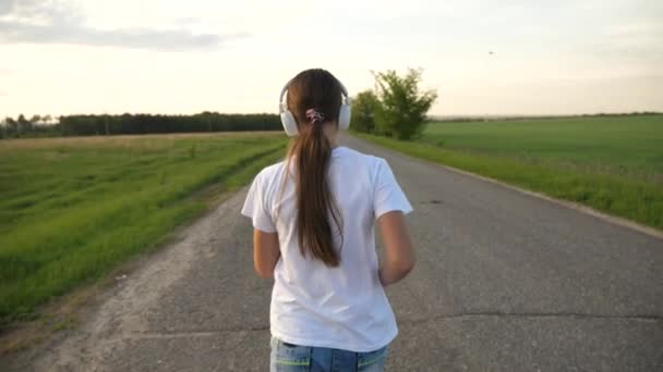 Dziewczynka jogging na drodze w godzinach wieczornych. dziewczyna idzie do uprawiania sportów i słucha muzyki w słuchawkach — Wideo stockowe