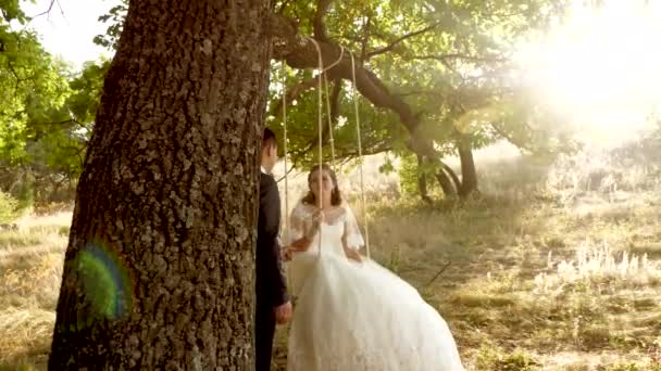 Щасливий наречений гойдається на гойдалках нареченої в красивій білій сукні в літньому парку. гойдалка на гілці дуба в літньому лісі і любляча пара — стокове відео