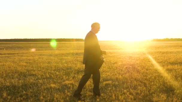 穿着严肃西装的老商人穿着黑色公文包, 在明亮的阳光下穿过田野, 参加一个重要的会议。慢动作 — 图库视频影像