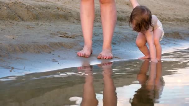 Kid plays on bank of the river, takes wet sand with his hand. mom with child walk on beach along the river. — Stock Video