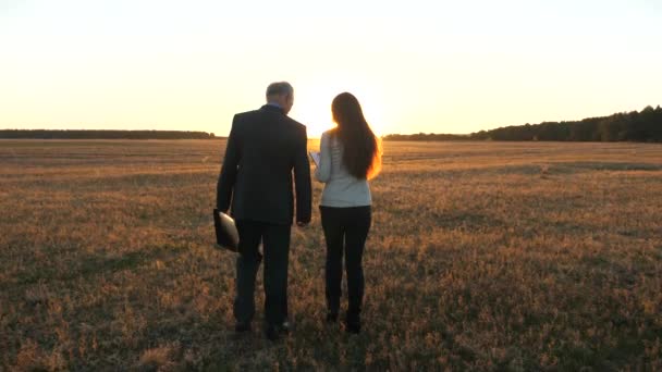 Hombre de negocios y mujer de negocios caminan a través del campo en los rayos del atardecer . — Vídeos de Stock