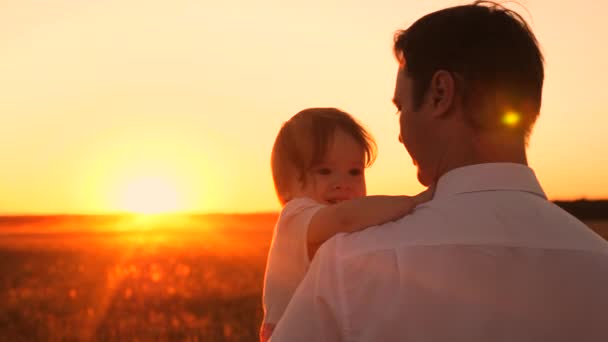 Papa trägt die glückliche kleine Tochter bei Sonnenuntergang auf dem Arm. Baby lächelt. Familienspaziergänge im Park am Abend in den Strahlen der roten Sonne. — Stockvideo