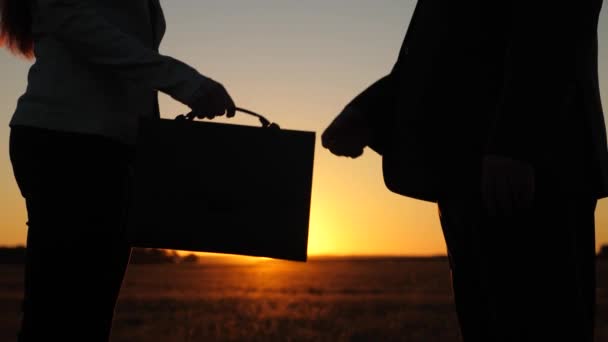 Uomo d'affari e giovane donna d'affari fatto affare, passare valigetta nera l'un l'altro e stringere la mano al tramonto sul campo. primo piano — Video Stock