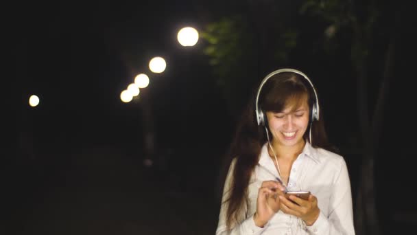 Dziewczyna idzie w nocy w parku miejskim w świetle latarni. Piękna dziewczyna w słuchawkach słucha muzyki na jej smartphone i tańce. Zwolnionym tempie — Wideo stockowe