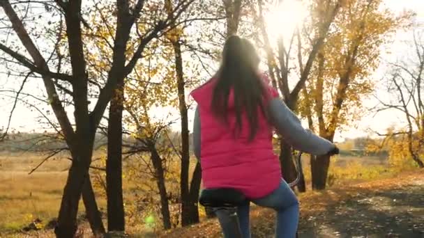 赤いジャケットの女の子は、沈む太陽の明るい光の通りに自転車を乗っています。黄色の木の背景。スローモーション. — ストック動画