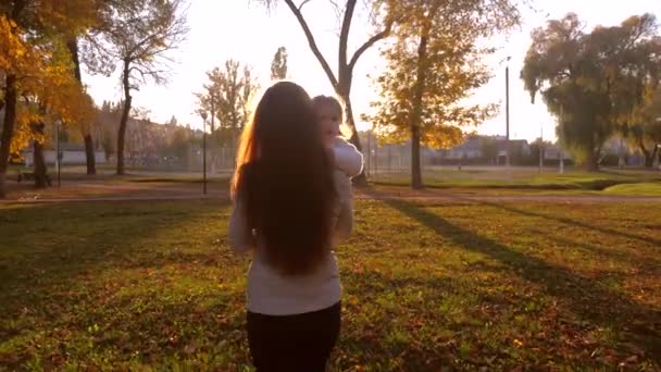 Mamá con un niño pequeño caminando en un parque de la ciudad iluminado por la sombra del sol brillante — Vídeos de Stock