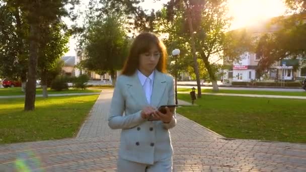 Девушка в светлом костюме идет на работу с планшетом в руке. Красивая деловая женщина гуляет по аллее в парке и проверяет электронную почту на планшете . — стоковое видео