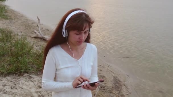 Девушка с наушниками ходить по песку рядом с водой и слушать музыку на планшете — стоковое видео