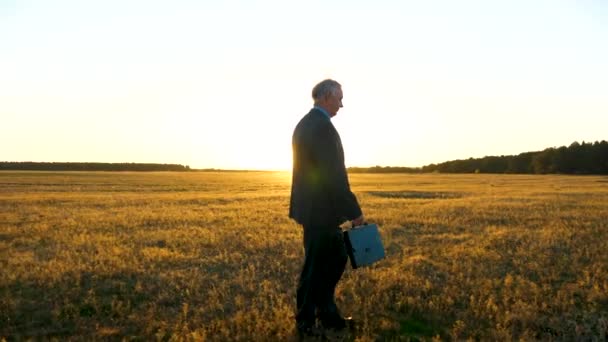 Um homem de negócios idoso de terno com pasta na mão caminha através do campo para aquecer raios do pôr do sol . — Vídeo de Stock